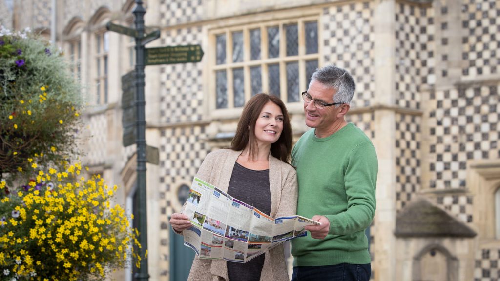A couple standing in front of King's Lynn Town Hall holding a Discover King's Lynn leaflet. The man is wearing a green jumper and the woman is wearing a beige cardigan.