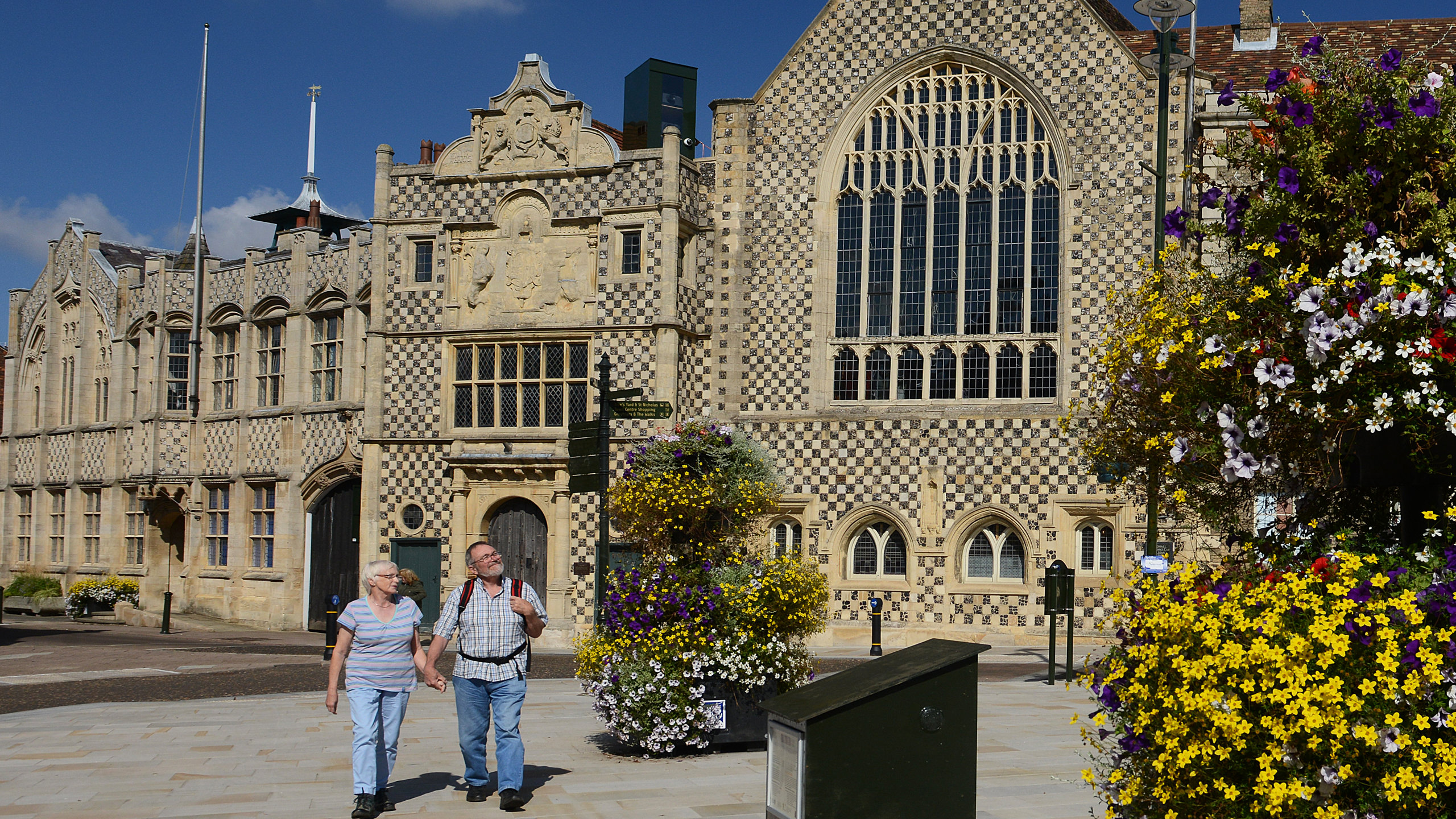 An image of an elderly couple walking away from King's Lynn Town Hall on Saturday Market Place. The town hall is a black and brown chequered building with large arched windows. The woman is wearing a striped t-shirt with jeans and the man is wearing a chequered shirt with a backpacked on his shoulders and wearing jeans.