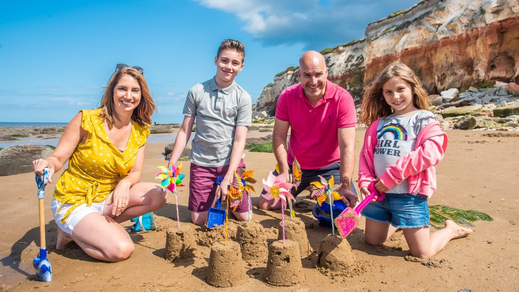 A family of four kneeling on Old Hunstanton beach, building sandcastles and smiling at the camera. From left to right, a woman is wearing a bright yellow top and white shorts holding a spade. Next to her sits a young boy wearing a button up grey polo shirt and purple shots. A man is sat beside him wearing a bright pink shirt and a young girl is beside him wearing a "Girl Power" t-shirt, pink jacket and denim shorts. The sandcastles have colourful flags in them and Old Hunstanton's famous red and white striped cliffs can be seen in the background.