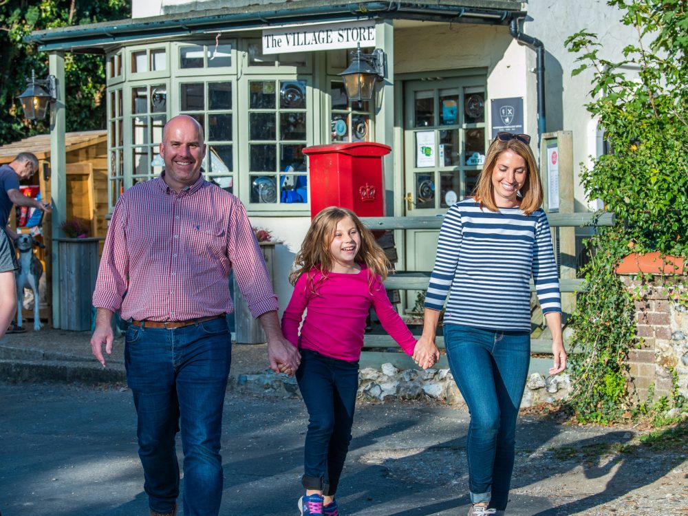 A happy shot of a family of four walking away from a village store. From left to right, a young boy is smiling at the camera wearing a short sleeved t-shirt and black jeans. An older man is standing in the middle of the image wearing a pink shirt and blue jeans. He is holding the hand of a little girl wearing a bright pink long sleeved t-shirt smiling. The little girl is also holding the hand of a woman who is smiling and wearing a striped long sleeved t-shirt and jeans.