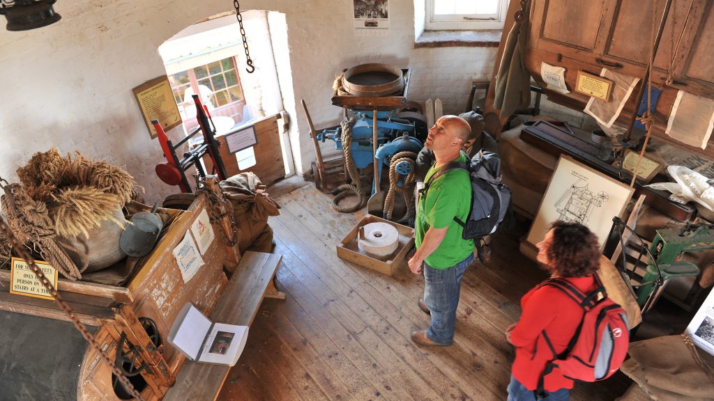 A couple walking around and looking at the exhibitions at Bircham Windmill. The man is wearing a green t-shirt with a back pack on his shoulders and the woman is wearing a red top, blue jeans and a red back pack.