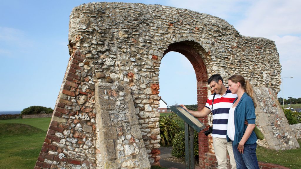 A young couple standing in front of the ruins of St Edmund's Chapel in Old Hunstanton. They are reading the information sign. The man is wearing a red and blue striped polo shirt and the woman is wearing a white top and blue cardigan. Both are smiling.