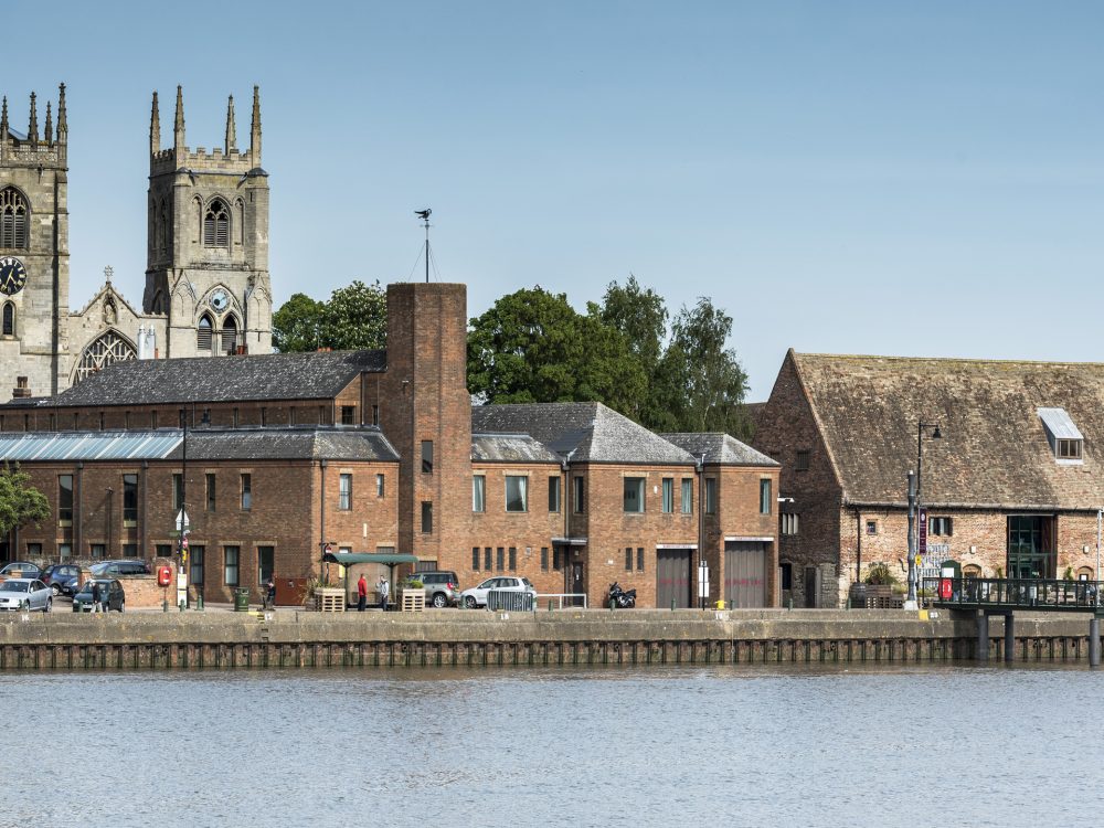 A long shot of South Quay in King's Lynn. Historic buildings including King's Lynn Minster, Clifton Tower and Marriotts Warehouse can be seen.