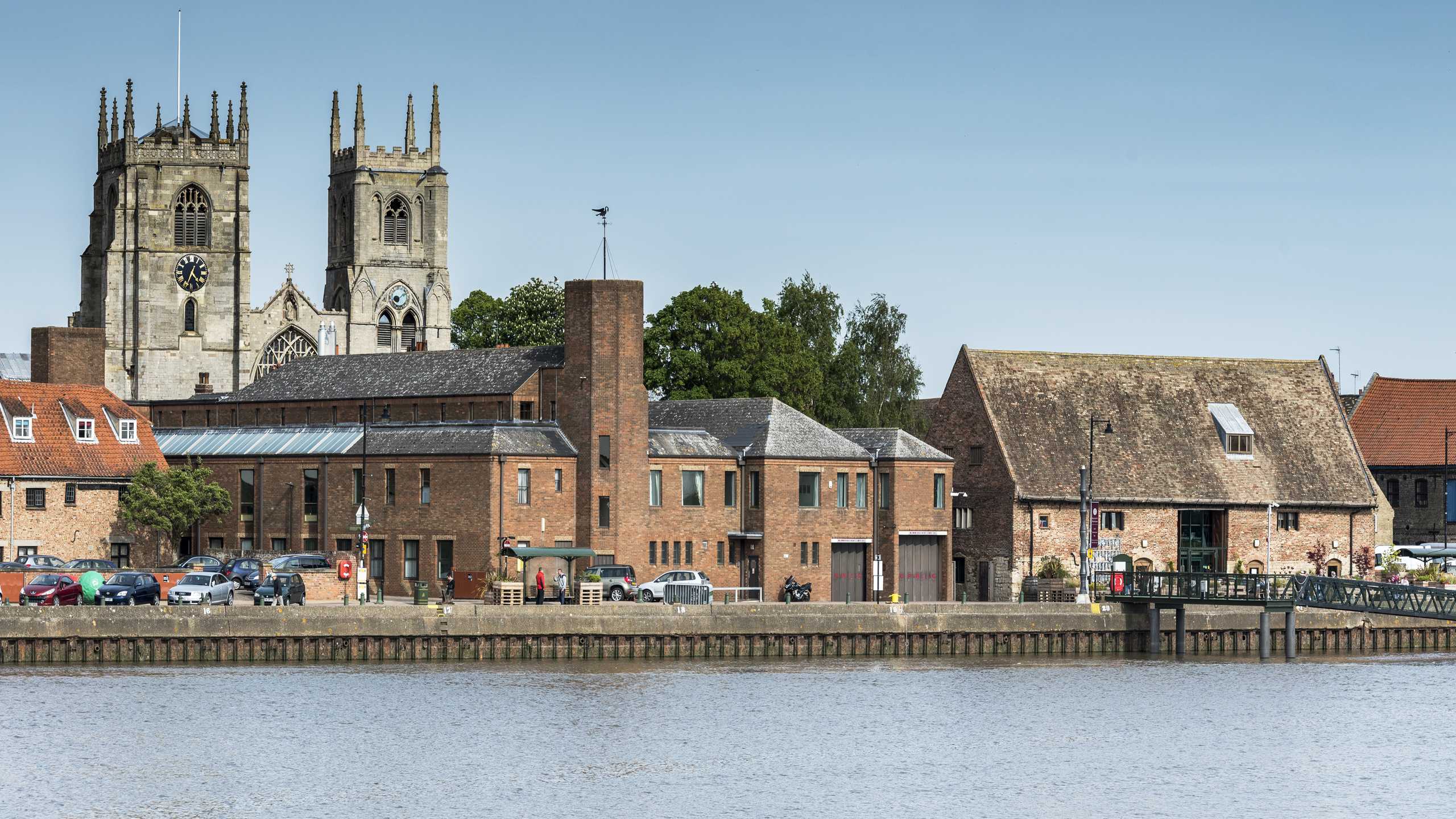 A long shot of South Quay in King's Lynn. Historic buildings including King's Lynn Minster, Clifton Tower and Marriotts Warehouse can be seen.