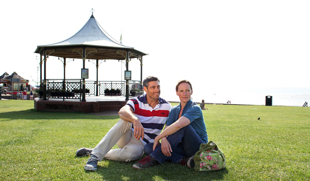 A young couple sitting in front of the bandstand on Hunstanton Green. From left to ring, the man is sat smiling into the distance wearing a striped polo shirt and beige trousers. The woman is sat beside him wearing a blue top and blue jeans.