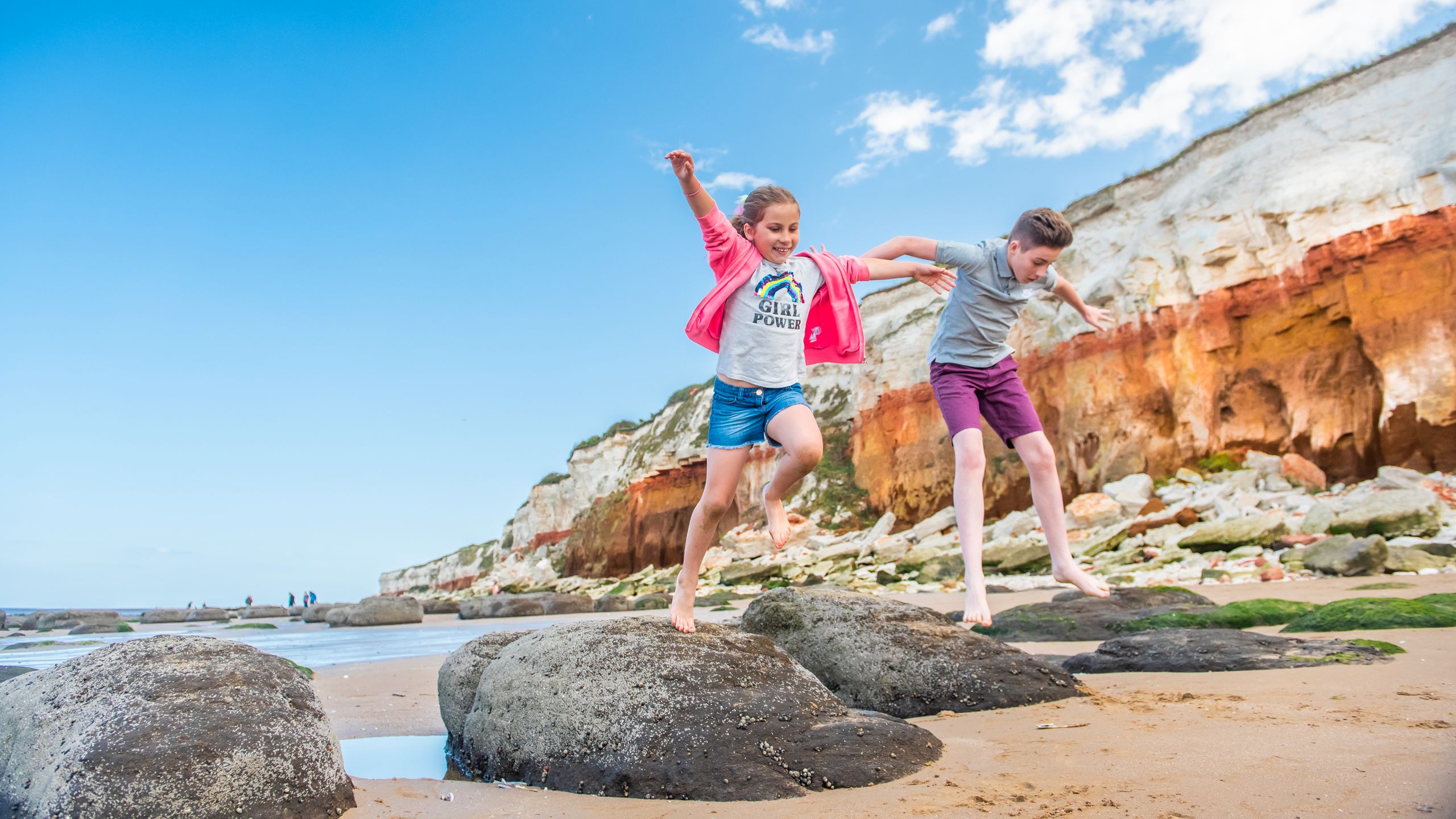 Two children jumping from rocks at Old Hunstanton beach. The girl is wearing a top that says 