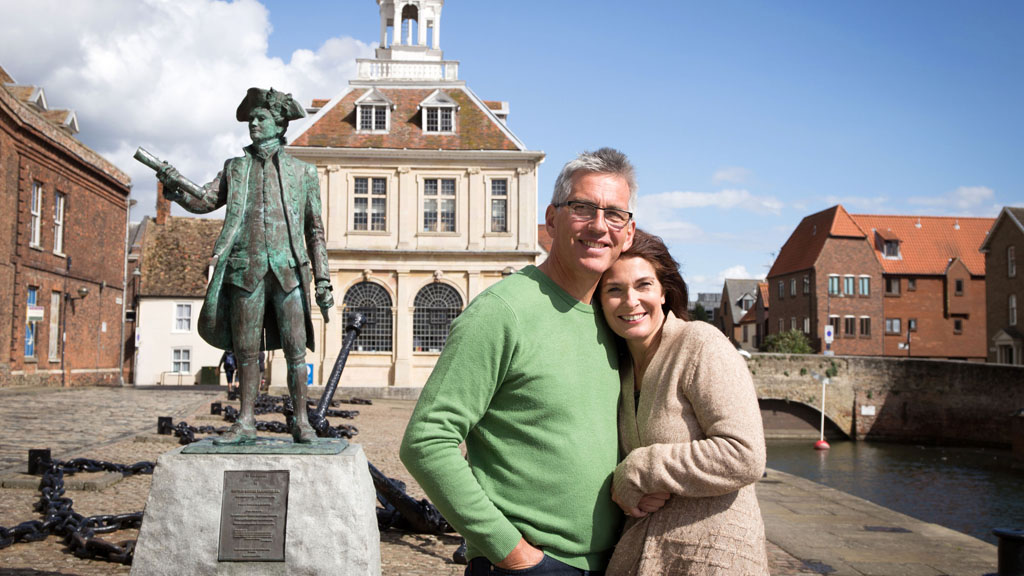 A couple standing in front of The Custom House and Captain Vancouver Statue at Purfleet Quay, King's Lynn. From left to right, the man is wearing a green jumper and the woman is wearing a beige cardigan.