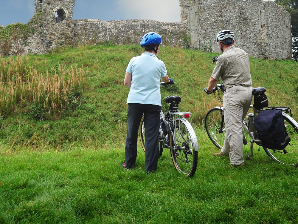 An elderly couple holding their bikes and looking over Castle Rising Castle.