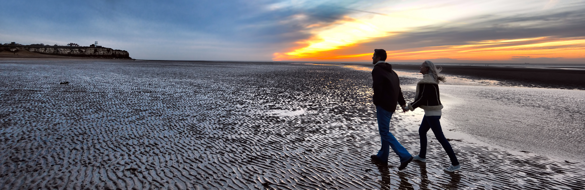 A panoramic shot of a couple holding hands on an empty beach at Old Hunstanton.