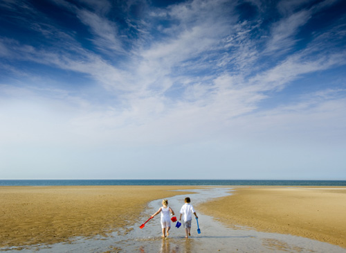 Two kids running towards the sea with buckets and spades, on a sunny day out at Holkham Beach in Norfolk