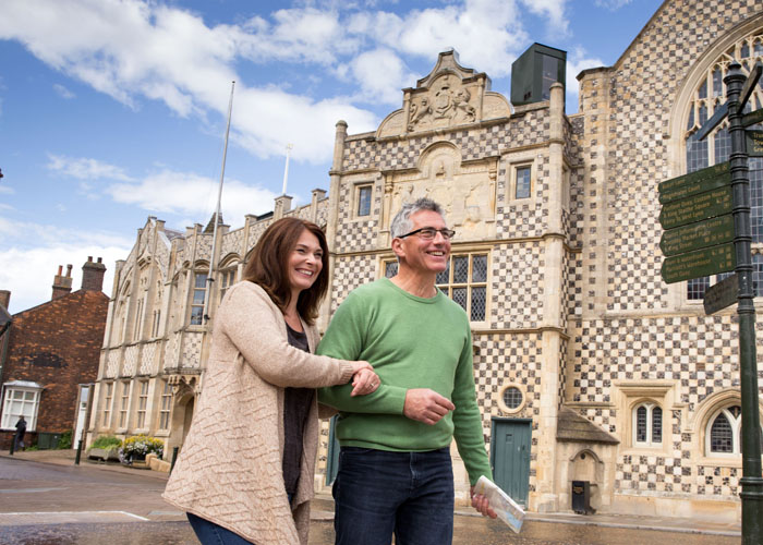 A couple standing in front of King's Lynn Town Hall. The man is wearing a green jumper and the woman is wearing a beige cardigan.