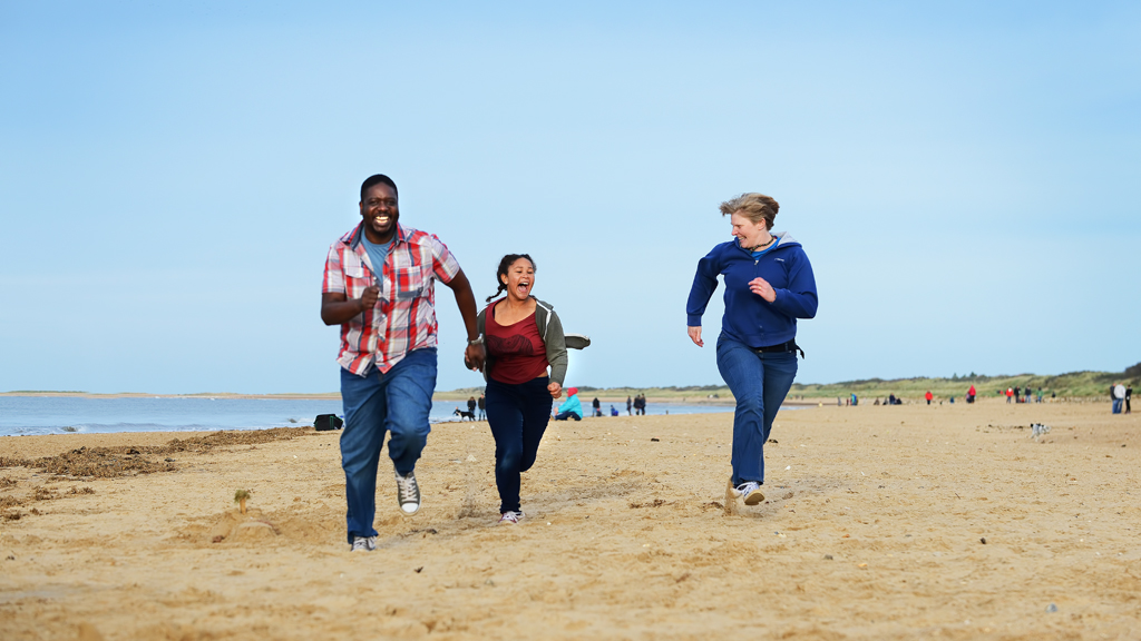 A family of three running down Hunstanton beach smiling and laughing, From left to right, a man is seen running whilst looking at his family smiling and is wearing a chequered shirt and blue jeans. A young girl is running in the middle smiling at the camera wearing a red top, grey jacket and dark blue jeans. On the right is a woman smiling at the camera wearing a dark blue top and darker blue hoodie and jeans.