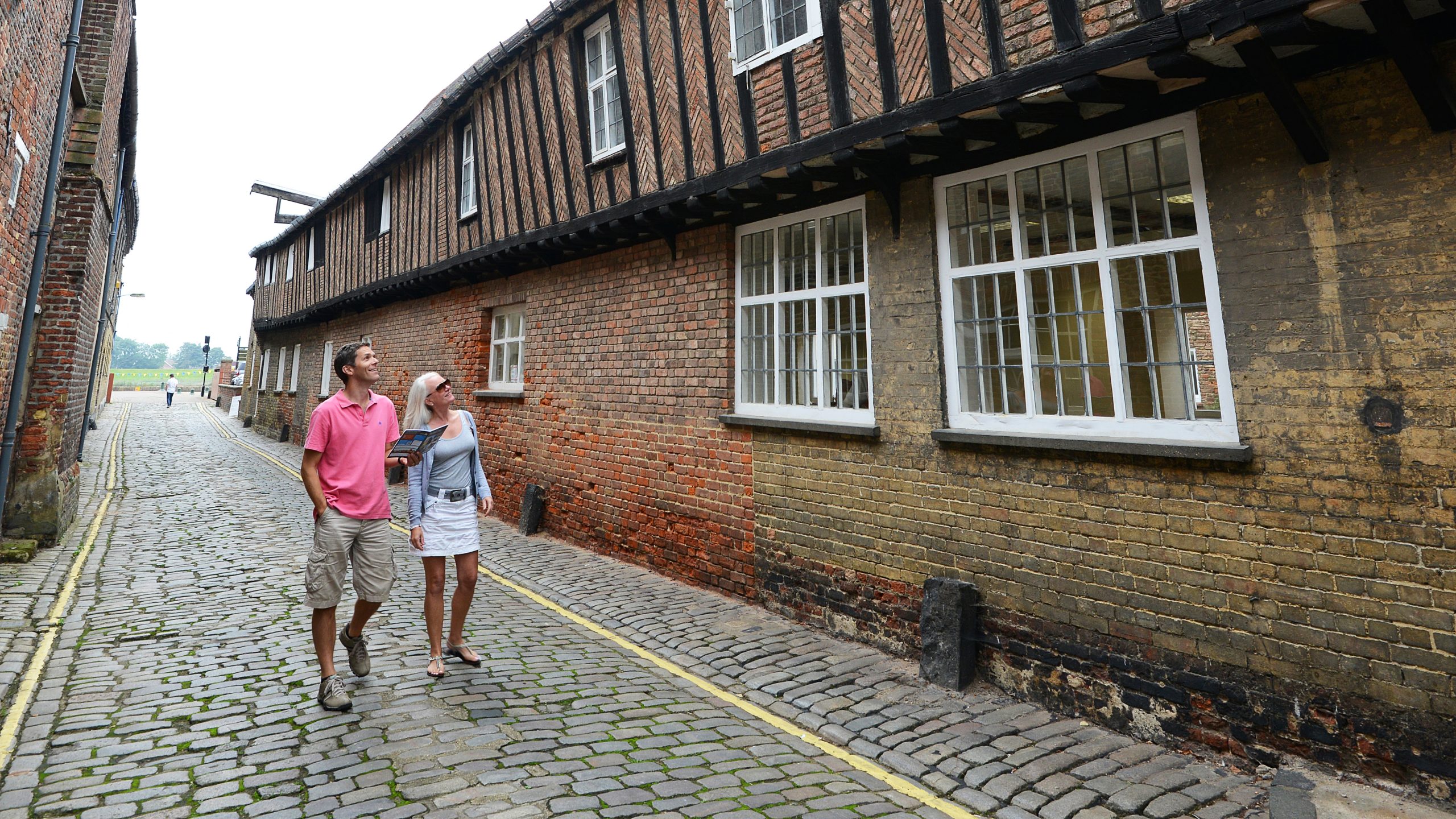 A panoramic shot of a young couple walking down a cobbled street next to historic Hanse House. The man is wearing a bright pink shirt, three quarter length shorts and trainers and the woman is wearing a light blue vest top, blue cardigan, white skirt and sandals. They are both looking up at Hanse House whilst holding a Visit West Norfolk leaflet.