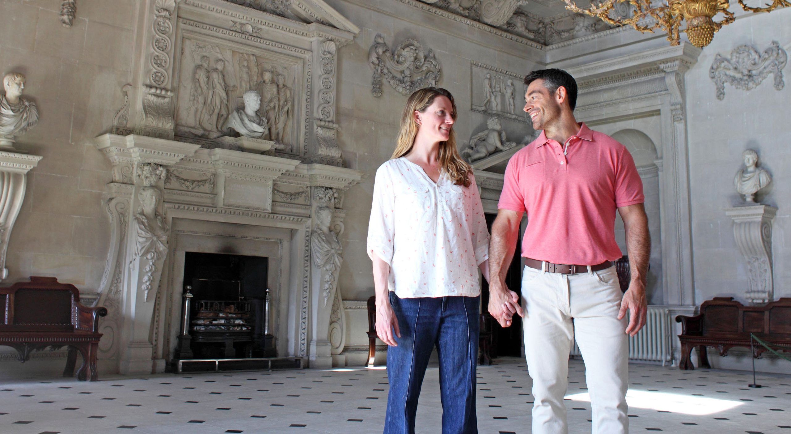 A couple standing in the Marble Room at Houghton Hall. The man is wearing a pink polo shirt and beige trousers and he is holding hands with a woman wearing a white top and blue jeans.