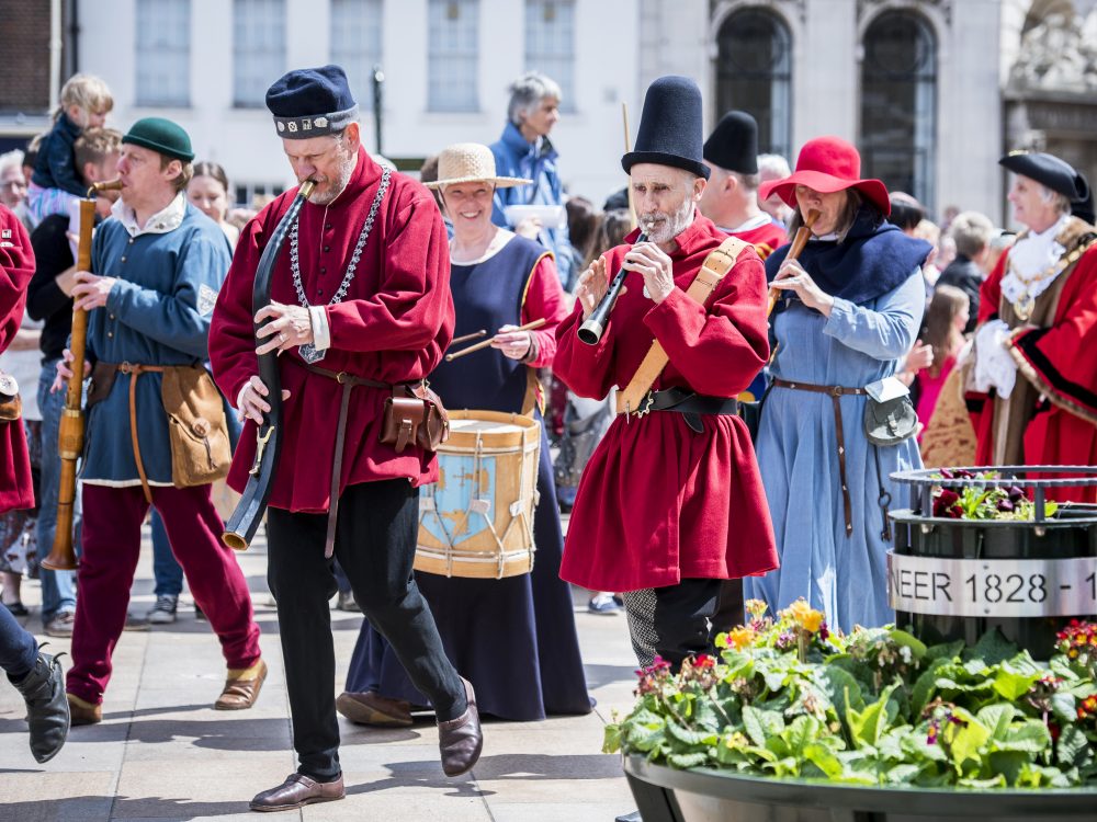 A happy shot of a family of four walking away from a village store. From left to right, a young boy is smiling at the camera wearing a short sleeved t-shirt and black jeans. An older man is standing in the middle of the image wearing a pink shirt and blue jeans. He is holding the hand of a little girl wearing a bright pink long sleeved t-shirt smiling. The little girl is also holding the hand of a woman who is smiling and wearing a striped long sleeved t-shirt and jeans.