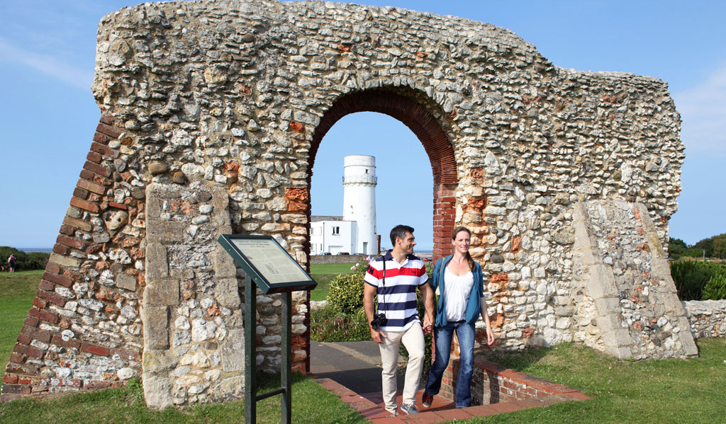 A young couple standing in front of the ruins of St Edmund's Chapel in Old Hunstanton. The man is wearing a red and blue striped polo shirt and the woman is wearing a white top and blue cardigan. Both are smiling.