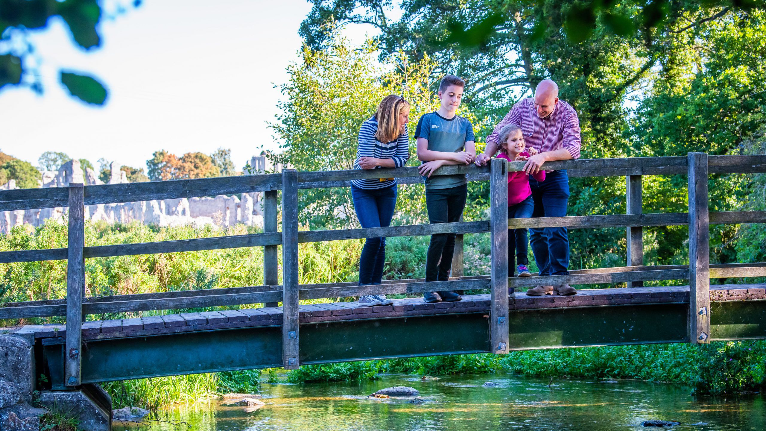 family of four standing on Castle Acre's wooden bridge. The ruins of Castle Acre Castle can be seen in the background.