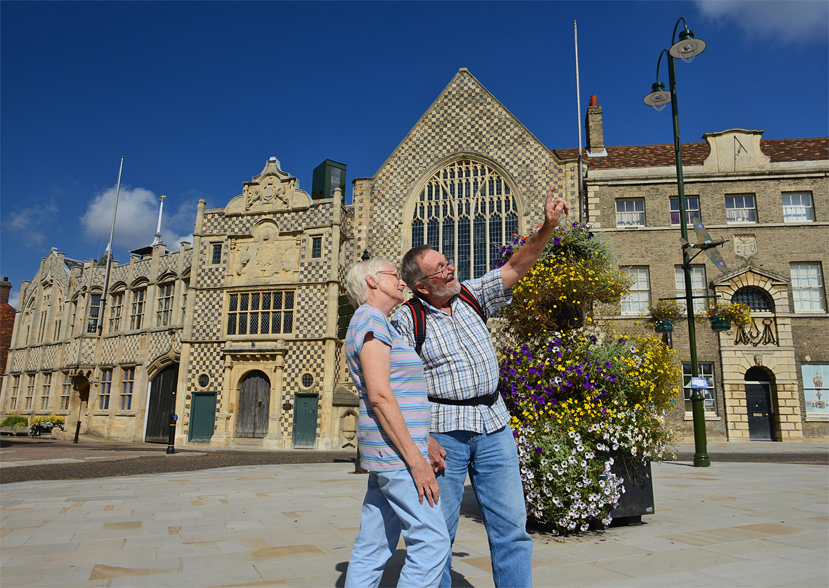 An elderly couple walking around Saturday Market Place. The woman is wearing a striped top and the man is wearing a chequered shirt with a backpack over his shoulder. King's Lynn's chequered town hall can be seen in the background.