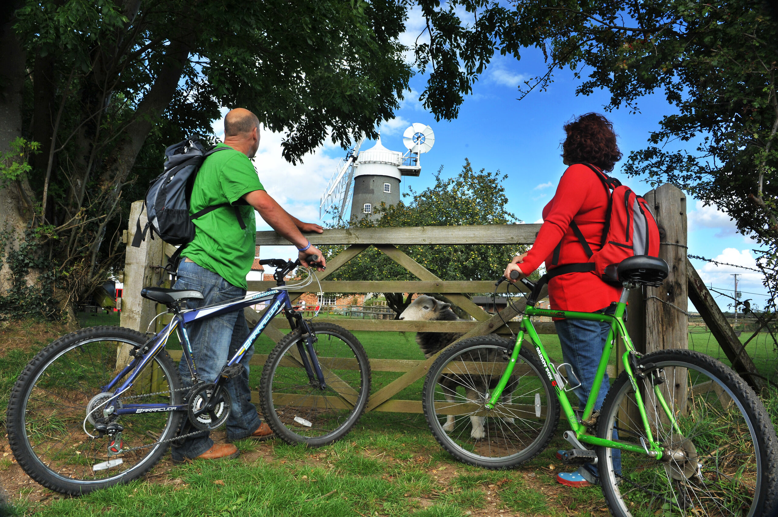 A couple holding their bikes up beside a gate looking at Bircham Windmill. The man is wearing a bright green t-shirt and the woman is wearing a long-sleeved red top.