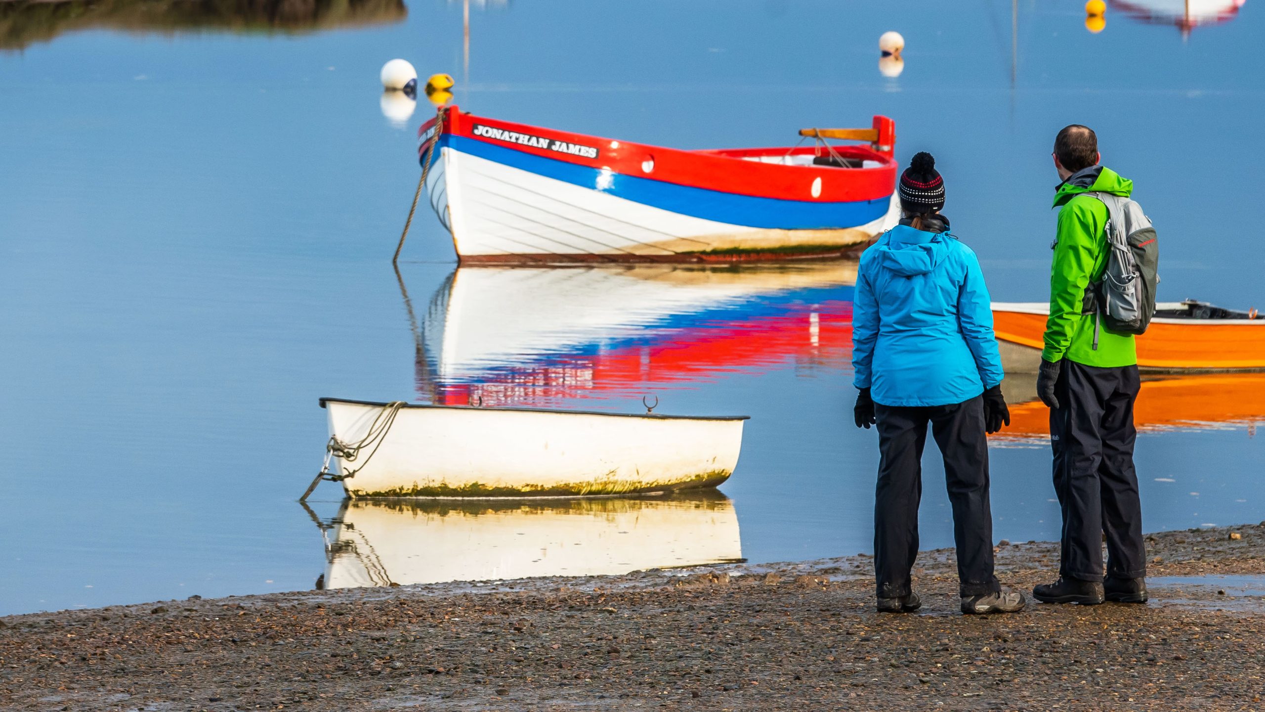 A couple looking at the colourful moored boats at Burnham Overy Staithe. The man is wearing a bright green jacket and backpack. The woman is wearing a woolly hat and bright blue jacket. The boats are painted white, red and blue.
