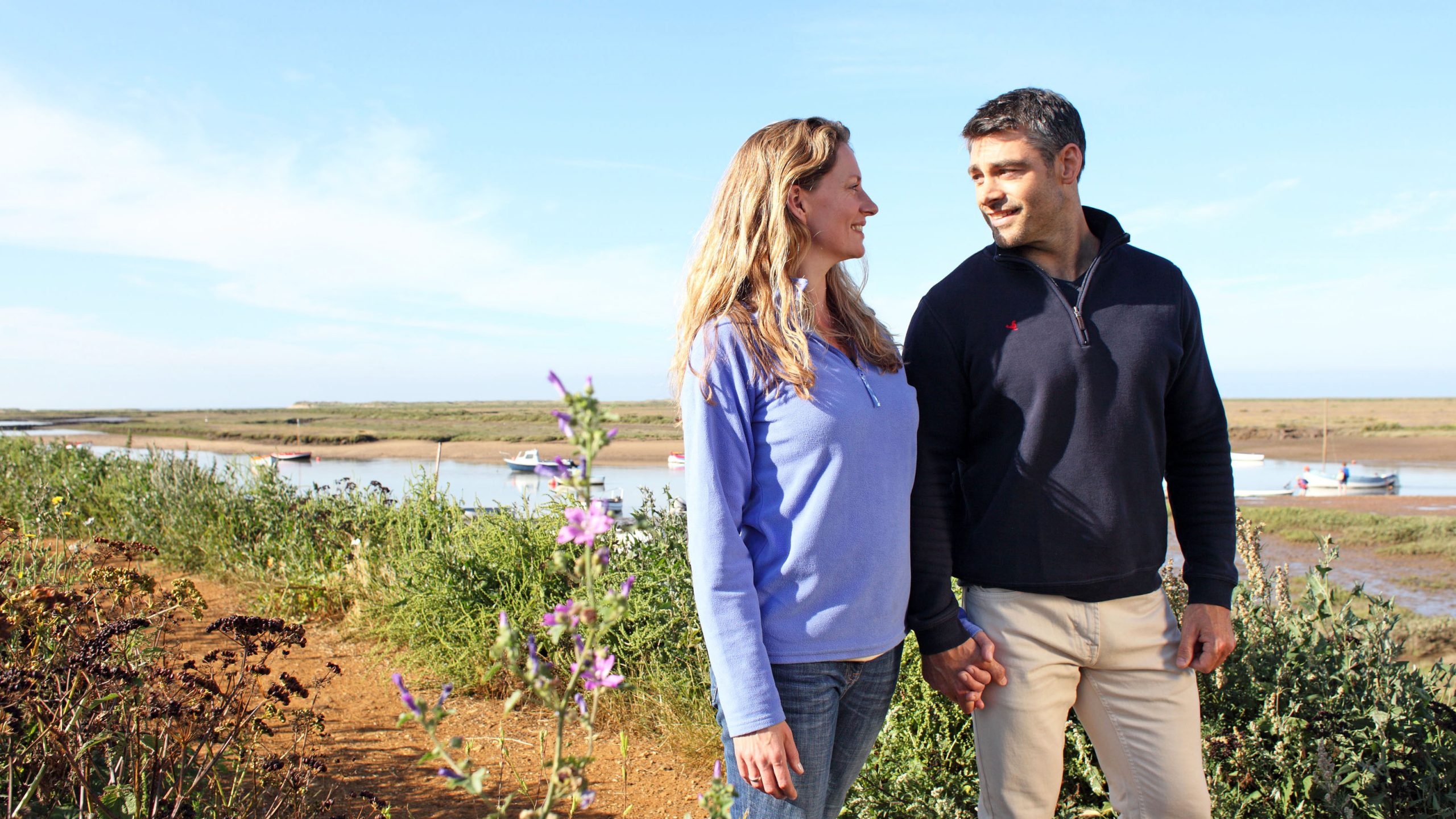 A couple smiling and walking along the pathway at Burnham Overy Staithe in West Norfolk.
