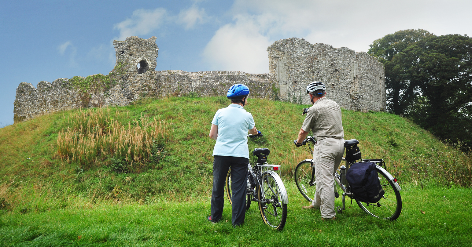 Man and woman holding their bikes in front of Castle Rising Castle, West Norfolk.