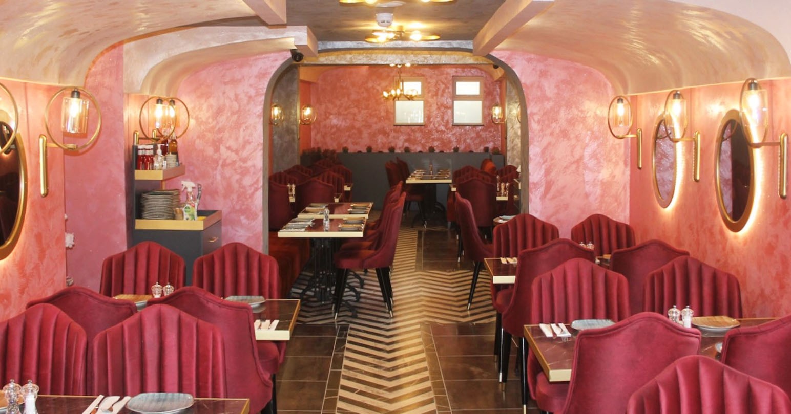 The pink interior of the Cave Kitchen, Downham Market. There is a walk way that separates the velvet shares and black and gold tables. Light-up mirrors are fixed on the pink walls.