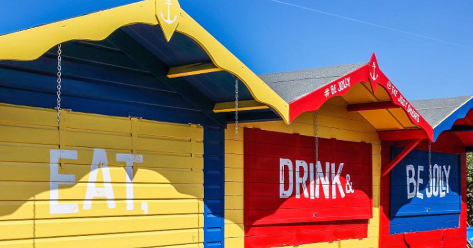 The three colourful red, yellow and blue huts at The Jolly Sailors. The huts are next to each other. The first hut has "Eat" painted on it". The second hut has "Drink &" painted on it. The third hut has "Be Jolly" painted on it.