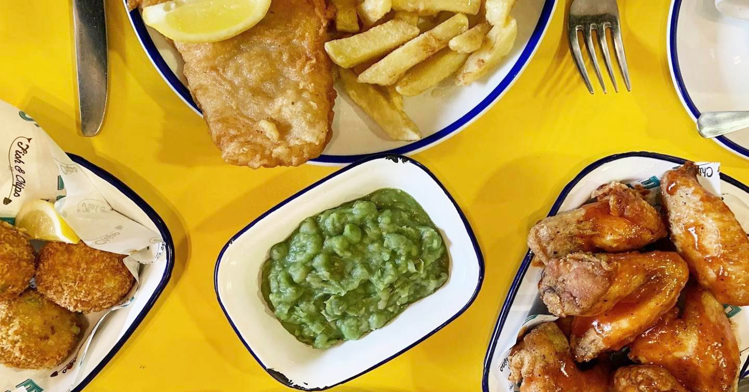 Dishes including Fish and Chips and Mushy Peas served on a yellow table at Eric's Fish and Chips in Thornham, West Norfolk.