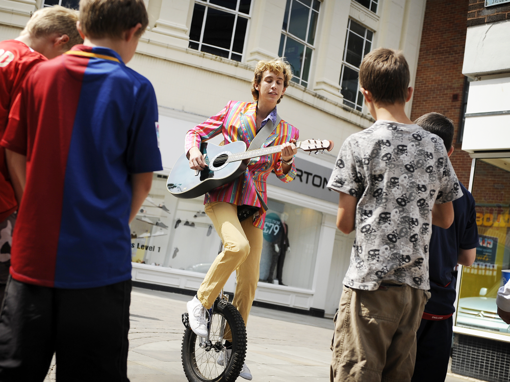 A performer playing guitar on a unicycle in front of a group of young children in King's Lynn, West Norfolk.