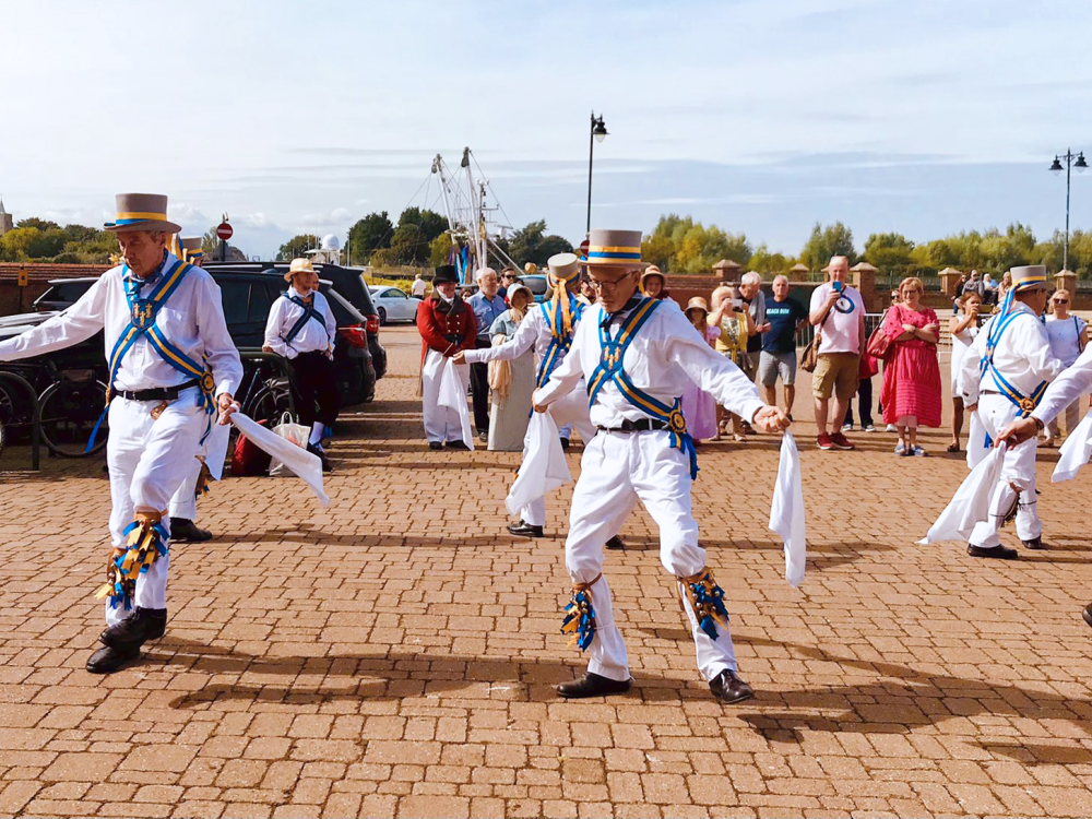 A happy shot of a family of four walking away from a village store. From left to right, a young boy is smiling at the camera wearing a short sleeved t-shirt and black jeans. An older man is standing in the middle of the image wearing a pink shirt and blue jeans. He is holding the hand of a little girl wearing a bright pink long sleeved t-shirt smiling. The little girl is also holding the hand of a woman who is smiling and wearing a striped long sleeved t-shirt and jeans.