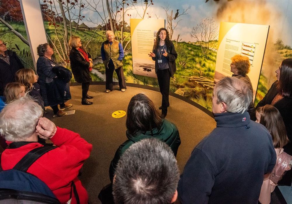 A happy shot of a family of four walking away from a village store. From left to right, a young boy is smiling at the camera wearing a short sleeved t-shirt and black jeans. An older man is standing in the middle of the image wearing a pink shirt and blue jeans. He is holding the hand of a little girl wearing a bright pink long sleeved t-shirt smiling. The little girl is also holding the hand of a woman who is smiling and wearing a striped long sleeved t-shirt and jeans.